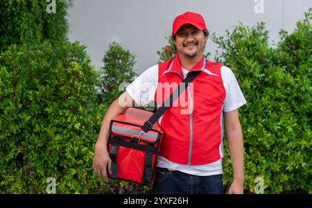 Sourire employé de livraison homme dans le chapeau rouge t-shirt uniforme thermique sac alimentaire sac à dos travail service de messagerie debout sur fond de nature Banque D'Images