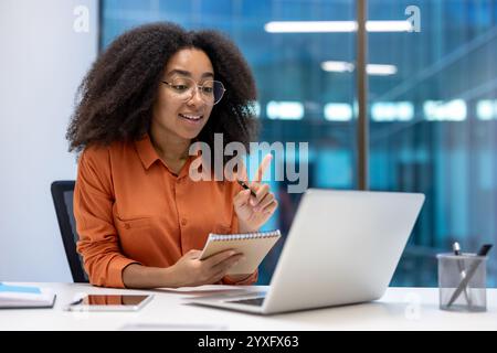 Un professionnel participe à une réunion virtuelle à l'aide de son ordinateur portable et note des notes dans son ordinateur portable. Elle est dans un cadre de bureau moderne avec de grandes fenêtres en verre Banque D'Images
