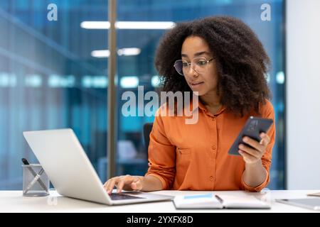 Femme confiante travaillant avec un ordinateur portable et un téléphone dans un espace de bureau contemporain. Elle est multitâche en se concentrant sur les deux appareils, mettant en valeur la productivité et le professionnalisme. Banque D'Images