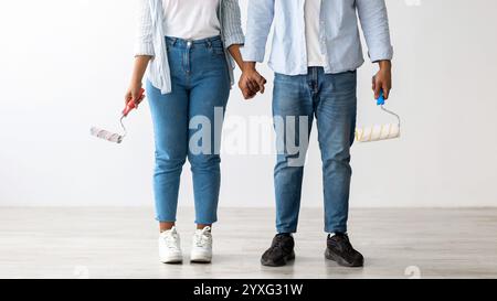 Rénovation, décoration et peinture. Couple afro-américain avec des rouleaux de peinture posant dans leur nouvel appartement, récolte Banque D'Images