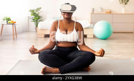 Femme noire de taille plus dans un casque VR, assise dans une posture lotus, méditant ou pratiquant le yoga à l'intérieur Banque D'Images