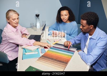 Équipe diversifiée de trois designers choisissant la couleur de la sellerie sur des échantillons de tissu tout en créant un design d'intérieur collaboratif assis à table au lieu de travail Banque D'Images