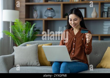Femme asiatique magasinant en ligne en utilisant un ordinateur portable et une carte de crédit, assis confortablement sur un canapé dans le cadre de la maison. Concept de commodité, de technologie et de commerce électronique dans la vie quotidienne. Banque D'Images