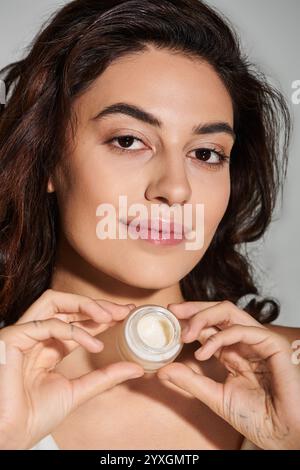 Belle jeune femme tient un produit de soin de la peau avec une expression rayonnante dans la lumière naturelle. Banque D'Images