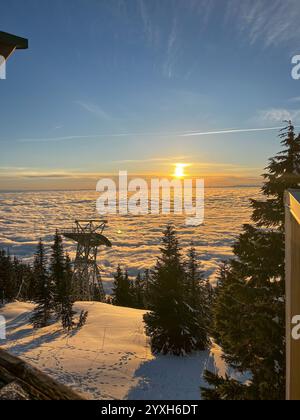 Vue sur le coucher du soleil depuis Grouse Mountain montrant la tour de remontée mécanique silhouette contre des nuages dorés en dessous, avec des pentes enneigées et des arbres à feuilles persistantes dans le f Banque D'Images