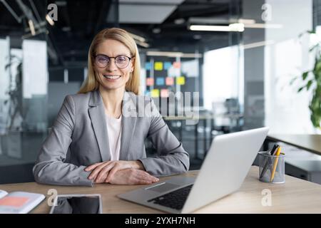 Femme d'affaires mature femme portant des lunettes et costume assis au bureau dans le bureau moderne. Souriante et confiante, elle travaille avec un ordinateur portable et un bloc-notes, mettant en valeur la productivité et le succès professionnels. Banque D'Images