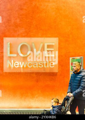Homme poussant une poussette avec un enfant devant un mur orange avec un panneau «LOVE Newcastle», soulignant la fierté locale et la vie urbaine Banque D'Images