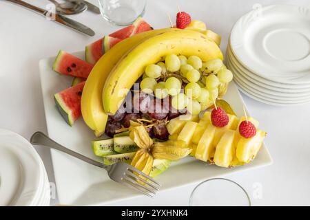 Ensemble de fruits frais sur une assiette avec bananes, raisins, ananas, kiwi, pastèque, framboises et ustensiles sur une table blanche, arrangement de fruits alimentaires i Banque D'Images