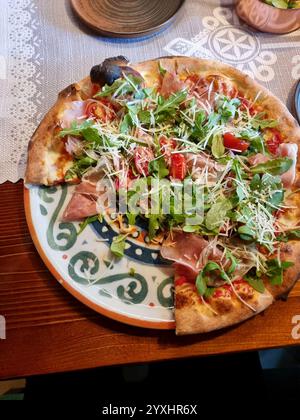 Pizza italienne premium garnie de roquette, prosciutto, tomates cerises et parmesan sur une assiette décorative, partiellement mangée, vue à la première personne, Banque D'Images