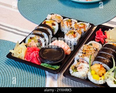 De nombreux rouleaux de sushi colorés différents avec sauce soja, wasabi, gingembre et garnitures dans un plateau noir sur une table décorative, commande de nourriture maison Banque D'Images