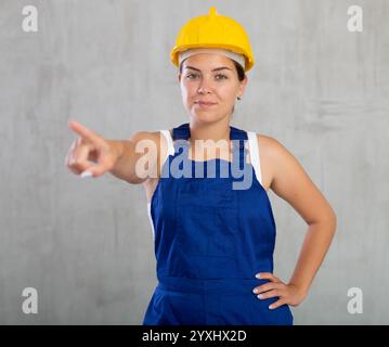 Travailleuse souriante en vêtements de travail et casque pointant quelque part Banque D'Images