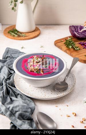 Soupe violette garnie de pacanes et de romarin dans un bol blanc sur une assiette mouchetée, accompagnée d'une cuillère et d'une serviette grise, avec une planche à découper et Banque D'Images