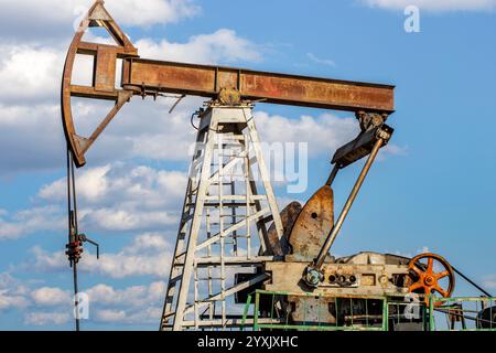 Un pommeau à huile altérée, utilisé pour extraire le pétrole, contre un ciel bleu vif avec des nuages dispersés. La structure métallique est rouillée, avec des engrenages visibles, Banque D'Images