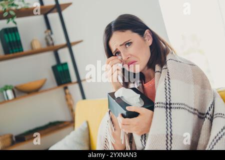 Femme émotionnelle dans le salon enveloppée dans une couverture, se sentant triste, tenant des tissus tout en étant assise sur un canapé dans un intérieur élégant de la maison Banque D'Images