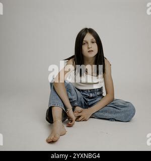 Fille aux cheveux bruns, 10 ans, assise pieds nus sur le sol. Prise de vue en studio Banque D'Images