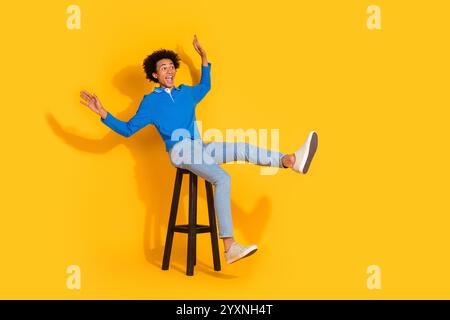 Photo du corps entier du jeune homme excité chaise en bois assis ludique regarder l'espace vide habillé vêtements bleus isolés sur fond de couleur jaune Banque D'Images