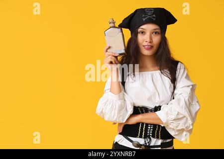 Belle jeune femme asiatique heureuse habillée en pirate avec une bouteille de rhum sur fond jaune Banque D'Images