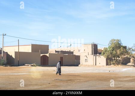 Un homme marche au soleil devant un village de Merzouga, au Maroc. Les constructions sont typiques des briques d'adobe et du ciment. L'homme porte un djell typique Banque D'Images