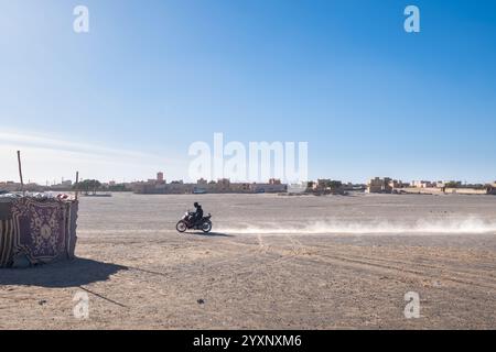 Une petite moto de type scooter traverse devant les hôtels du désert de Merzouga au Maroc. Le terrain est aride et sec et la moto monte Banque D'Images
