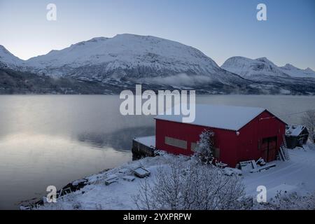 Cabine rouge traditionnelle sur la côte d'un fjord (mer de Norvège). Majestueuse montagne couverte de neige en arrière-plan. Ciel lumineux. Tennevoll, Norvège. Banque D'Images