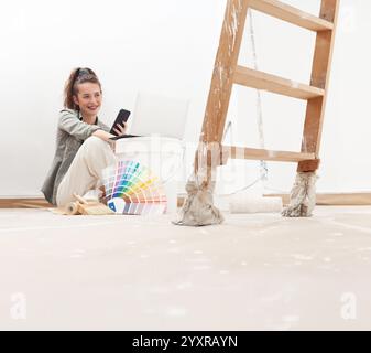 Femme heureuse utilisant son téléphone et son ordinateur portable pour chercher et acheter de la peinture murale pendant sa rénovation de maison. Elle est assise près d'un seau de peinture et d'un swa de couleur Banque D'Images