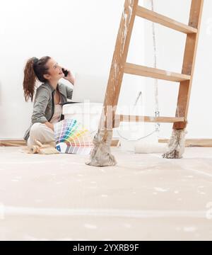 Femme heureuse utilisant son téléphone et son ordinateur portable pour chercher et acheter de la peinture murale pendant sa rénovation de maison. Elle est assise près d'un seau de peinture et d'un swa de couleur Banque D'Images