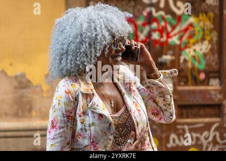 Élégante, afro-américaine dame avec des cheveux afro hoary et des lunettes de soleil parlant sur téléphone portable dans le centre-ville. Concept : pro-vieillissement, communication Banque D'Images