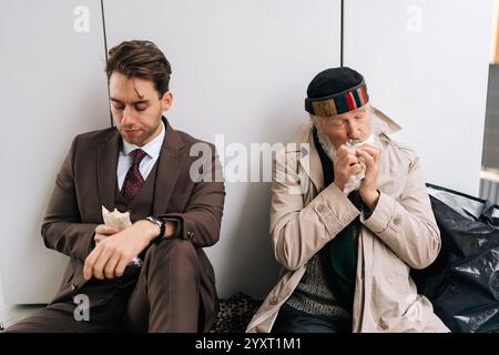 Homme d'affaires triste en costume et homme sans-abri senior affamé assis sur un morceau de carton sur la rue de la ville, manger des sandwichs et partager le moment Banque D'Images