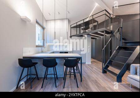 Un appartement loft élégant et contemporain avec une cuisine ouverte avec tabourets de bar, un espace de travail en mezzanine et un design minimaliste épuré. Moderne Banque D'Images