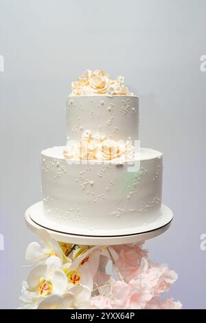 Un gâteau de mariage à deux niveaux magnifiquement décoré présente un délicat glaçage blanc et des accents floraux complexes sur le dessus. Il est affiché sur un simple stand, en Banque D'Images