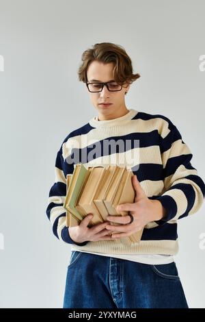 Un jeune homme dans un pull rayé se tient réfléchi, tenant une pile de livres dans un studio. Banque D'Images