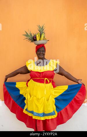 Une femme Palenquera portant Une robe rouge, jaune et bleue, tenant sa jupe avec Un panier de fruits équilibré sur sa tête Banque D'Images