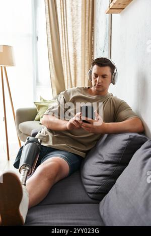 Beau jeune homme est assis confortablement sur un canapé, concentré sur son smartphone, portant des écouteurs. Banque D'Images