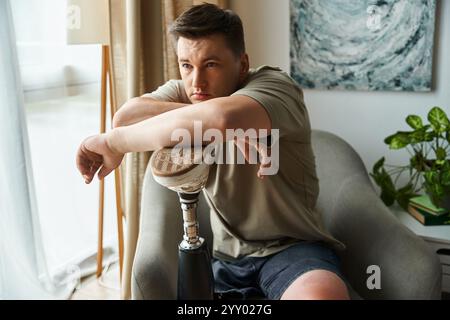 Jeune homme avec une jambe prothétique se détend tout en étant profondément dans la pensée dans une pièce lumineuse. Banque D'Images
