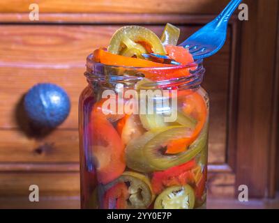 Gros plan d'une fourchette soulevant et égouttant quelques piments jalapeno rouges et verts tranchés, marinés dans du vinaigre d'alcool, d'un bocal en verre. Banque D'Images