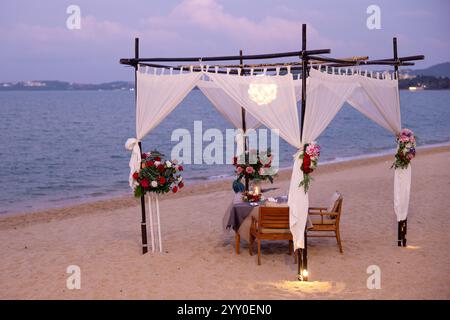 Élégante configuration de plage avec une table à manger sous un auvent blanc orné de fleurs, sur fond d'océan calme, parfait pour une GE romantique Banque D'Images