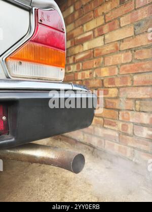 Gros plan des fumées du tuyau d'échappement arrière d'une vieille voiture vintage classique à moteur à essence, debout dans un garage en briques. Banque D'Images