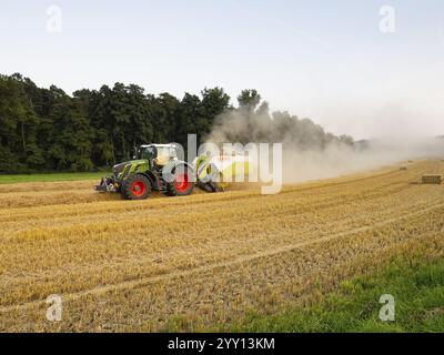 Agriculteur pressant la paille avec son tracteur et sa presse sur un champ après la récolte de maïs, l'air au-dessus du paysage couvert de poussière de la paille sèche, Nort Banque D'Images