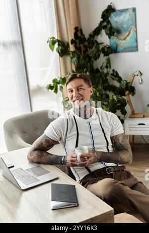 Beau jeune homme avec des tatouages est assis confortablement à un bureau tenant une tasse de café. Banque D'Images