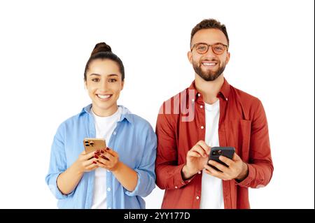 Homme et femme joyeux tenant des smartphones, paraissant heureux tout en s'engageant dans du contenu numérique, en envoyant des messages avec des amis, vêtus de façon décontractée, debout sur un fond blanc isolé, souriant à la caméra Banque D'Images