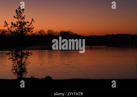 Sur le fond d'un lac profond, un coucher de soleil orange éclate dans le ciel, des vagues de lumière à la surface de l'eau reflètent des couleurs chaudes, silhouettes Banque D'Images