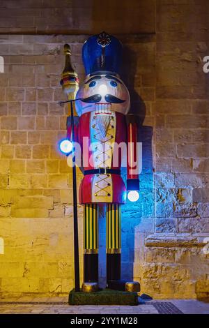 Soldat casse-noix géant en bois avec une tenue rouge et jaune vif, illuminé par des lumières bleues colorées et chaudes, se tient contre un mur de pierre texturé Banque D'Images