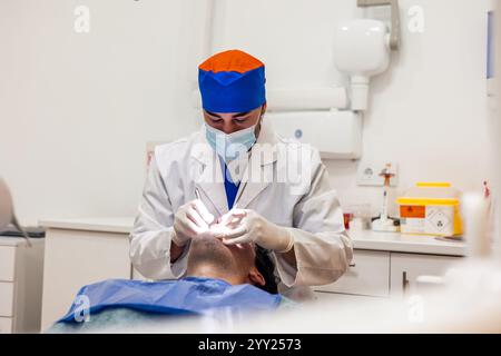 Dentiste portant un masque chirurgical et des gants effectuant un examen dentaire sur un patient masculin allongé sur un fauteuil dentaire dans une clinique moderne, en utilisant des instruments dentaires Banque D'Images