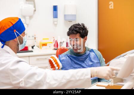 Un dentiste portant un masque et des gants montre une prothèse dentaire à un patient masculin qui rit dans une clinique dentaire, favorisant la santé buccodentaire et les soins dentaires Banque D'Images