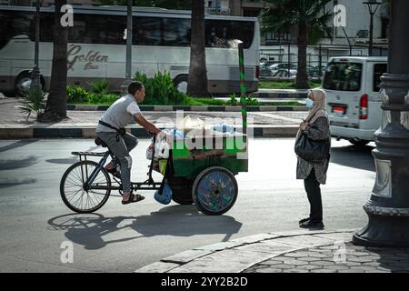 Une scène de rue à Alexandrie, Egypte. Un homme fait du vélo avec un chariot mobile équipé d'une cuisinière, de sacs et de fournitures, tandis qu'une femme se trouve à proximité. Banque D'Images