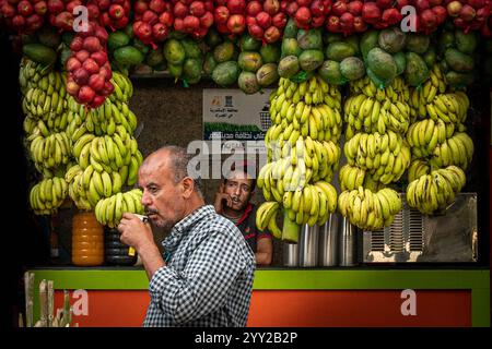 Un stand de jus vibrant à Alexandrie, en Égypte, avec des bananes suspendues, des pommes, des avocats. Un homme sirote du jus pendant qu'un vendeur attend au milieu des étalages de fruits colorés Banque D'Images