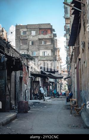 Ruelle étroite à Alexandrie, en Égypte, bordée de vieux bâtiments, de boutiques de rue et de résidents se détendant sur des chaises, capturant la vie urbaine égyptienne. Banque D'Images