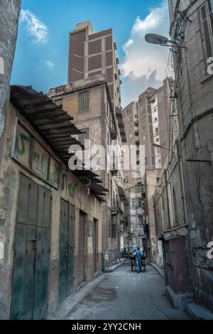 Ruelle étroite à Alexandrie, en Égypte, entourée de bâtiments altérés, de murs de graffitis et d'une vie urbaine animée avec des habitants poussant des chariots sous un ciel Banque D'Images