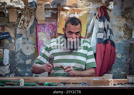 Un artisan dans une chemise verte rayée travaille intensément avec des outils dans un atelier rempli de bois fait à la main et de murs rustiques à Alexandrie, en Égypte. Banque D'Images