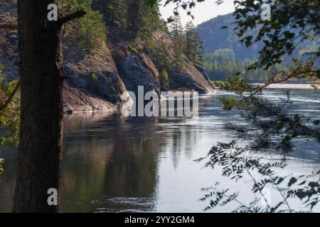 Une scène sereine au bord de la rivière encadrée par des arbres, avec des eaux calmes reflétant des falaises rocheuses et des forêts verdoyantes sous la lumière douce du soleil. Banque D'Images